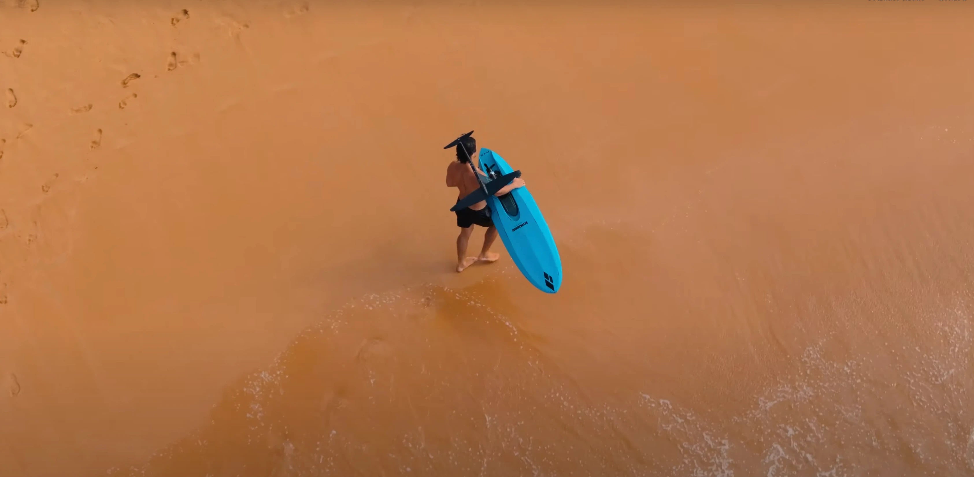 Individual carrying a blue foilboard on a sandy beach, showcasing Australian made Hydrofoil Boards from Amos Shapes.