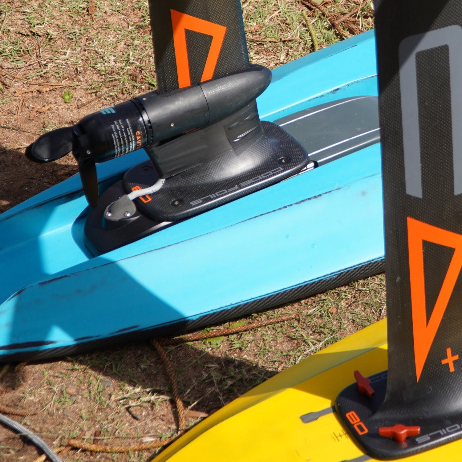 Close up shot of a Foil Drive assist kit mounted on a blue Kruzer RTS carbon foilboard by Amos Shapes on the beach