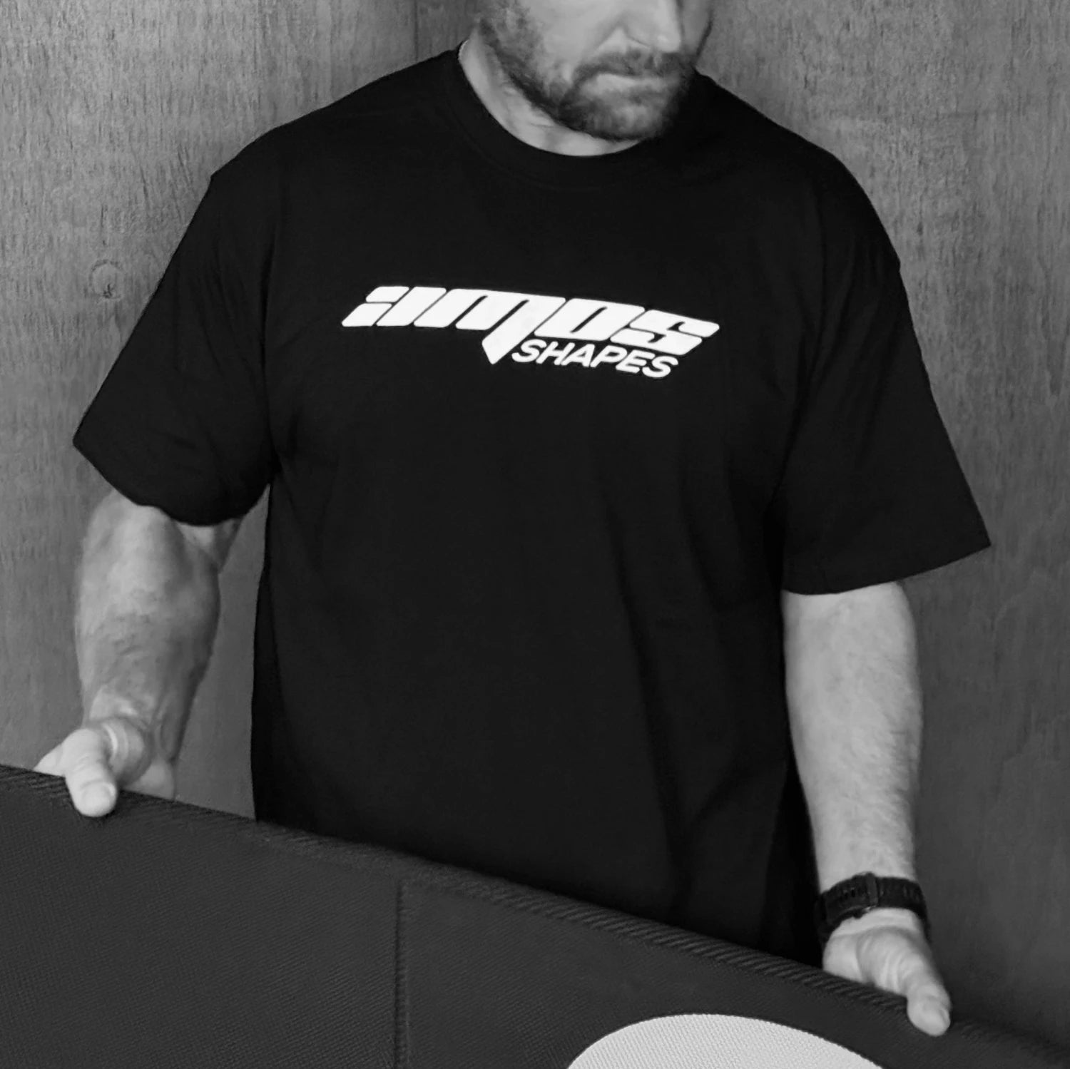 A foilboard shaper holding an Amosh Shapes foilboard and wearing a black Amos Shapes tee-shirt with the white Amos Shapes logo on the front