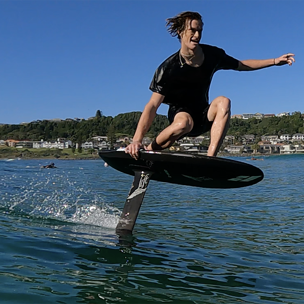 Team rider Luca smiling while riding a black Gen-Z carbon foilboard by Amos Shapes mounted with a Lift Foils hydrofoil on it in ocean conditions on a sunny day